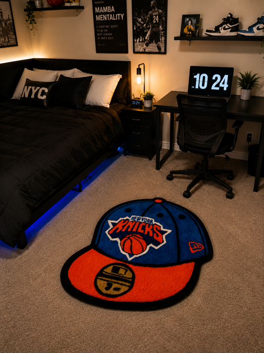 A New York Knicks themed shaped rug on a beige carpet in a modern bedroom setting. The rug is designed to look like a New Era 501 baseball cap, featuring the team's blue crown with orange and white 'New York Knicks' text and a basketball graphic, and a bright orange front brim with a gold and black New Era logo.
